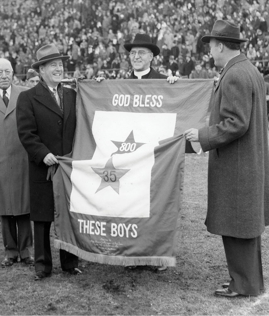 Vintage black and white photo of Father Flanagan and two other men holding a cloth banner that reads 'God Bless These Boys'.